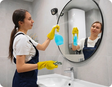 Kobieta stoi przed umywalką, widziana z profilu, trzymając w jednej ręce niebieski rozpylacz do środków czyszczących, a w drugiej – szmatkę lub serwetkę. Ma na sobie jaskrawożółte gumowe rękawiczki, białą koszulkę i ciemny strój roboczy, być może fartuch lub kombinezon do sprzątania. Jej postawa jest naturalna, jest lekko pochylona do przodu, skupiona na swoim zadaniu, wzrok skierowany na lustro lub w bok.